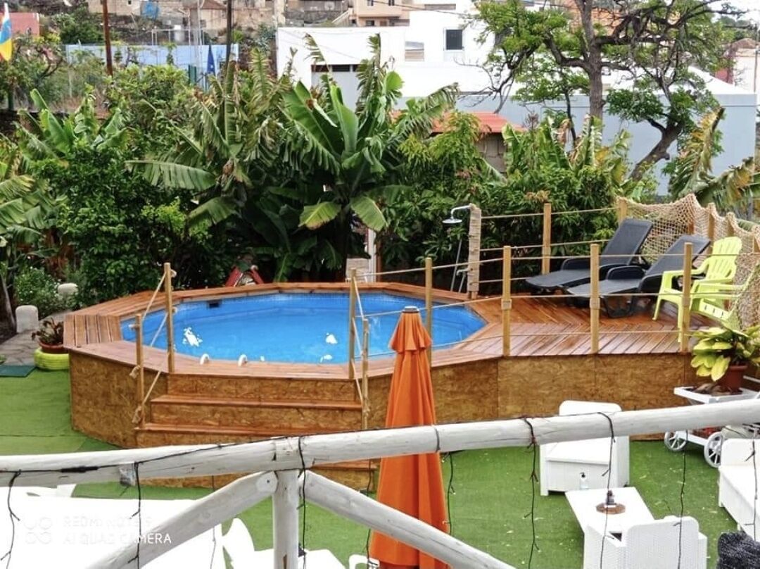 Un patio trasero con piscina de madera, rodeado de abundante vegetación y una terraza de madera.