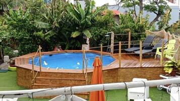 Un patio trasero con piscina de madera, rodeado de abundante vegetación y una terraza de madera.