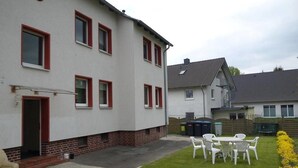 Il s’agit d’un immeuble résidentiel de deux étages, à l’extérieur blanc et aux encadrements de fenêtres rouges. On y trouve un petit patio avec une table et des chaises, ainsi qu’un jardin avec des fleurs jaunes.