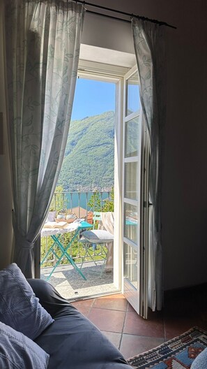 A room with a bed, a view of a balcony with outdoor furniture, and a mountainous landscape.