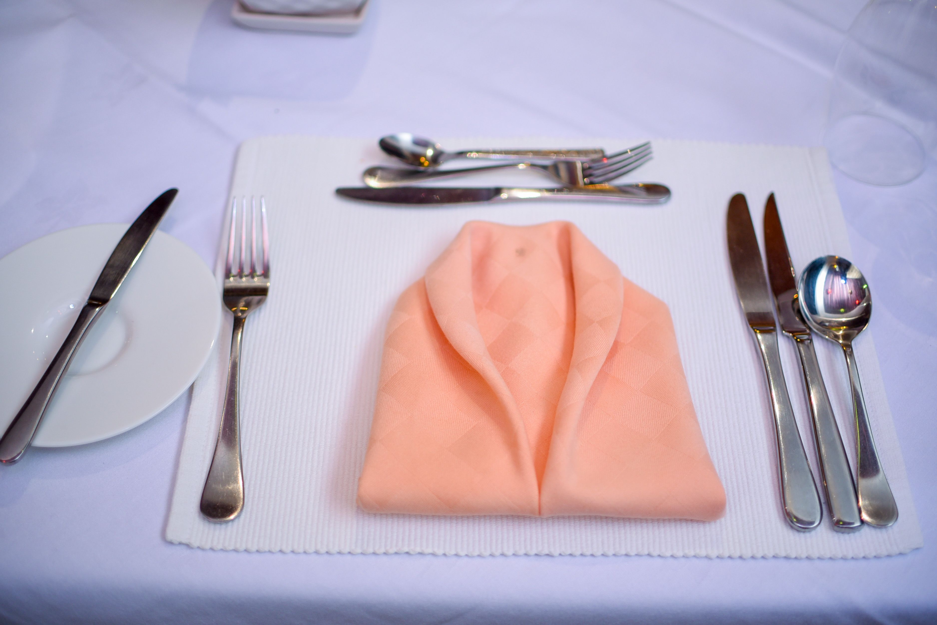Una mesa bien dispuesta con servilleta doblada, cubiertos y un plato sobre un camino de mesa blanco.