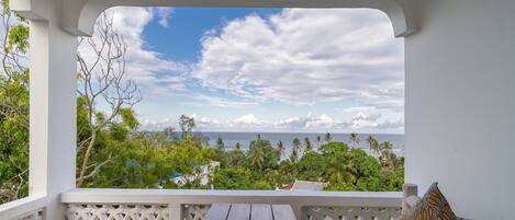A balcony with a view of the ocean, a wooden table, and a sofa with cushions.