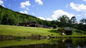 Un chalet comprenant un grand bâtiment et une petite cabane, entourés d’une colline verdoyante et d’un étang.