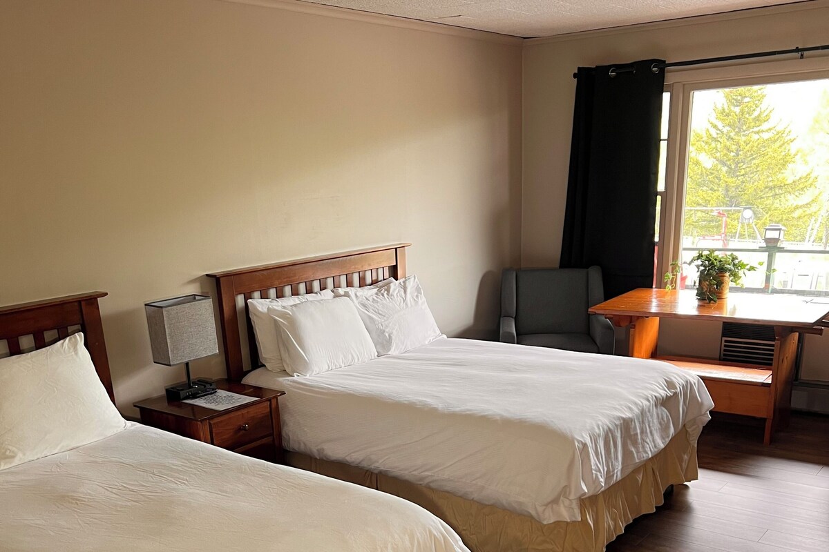 A hotel room with two beds, a desk, and a window with a view of trees.