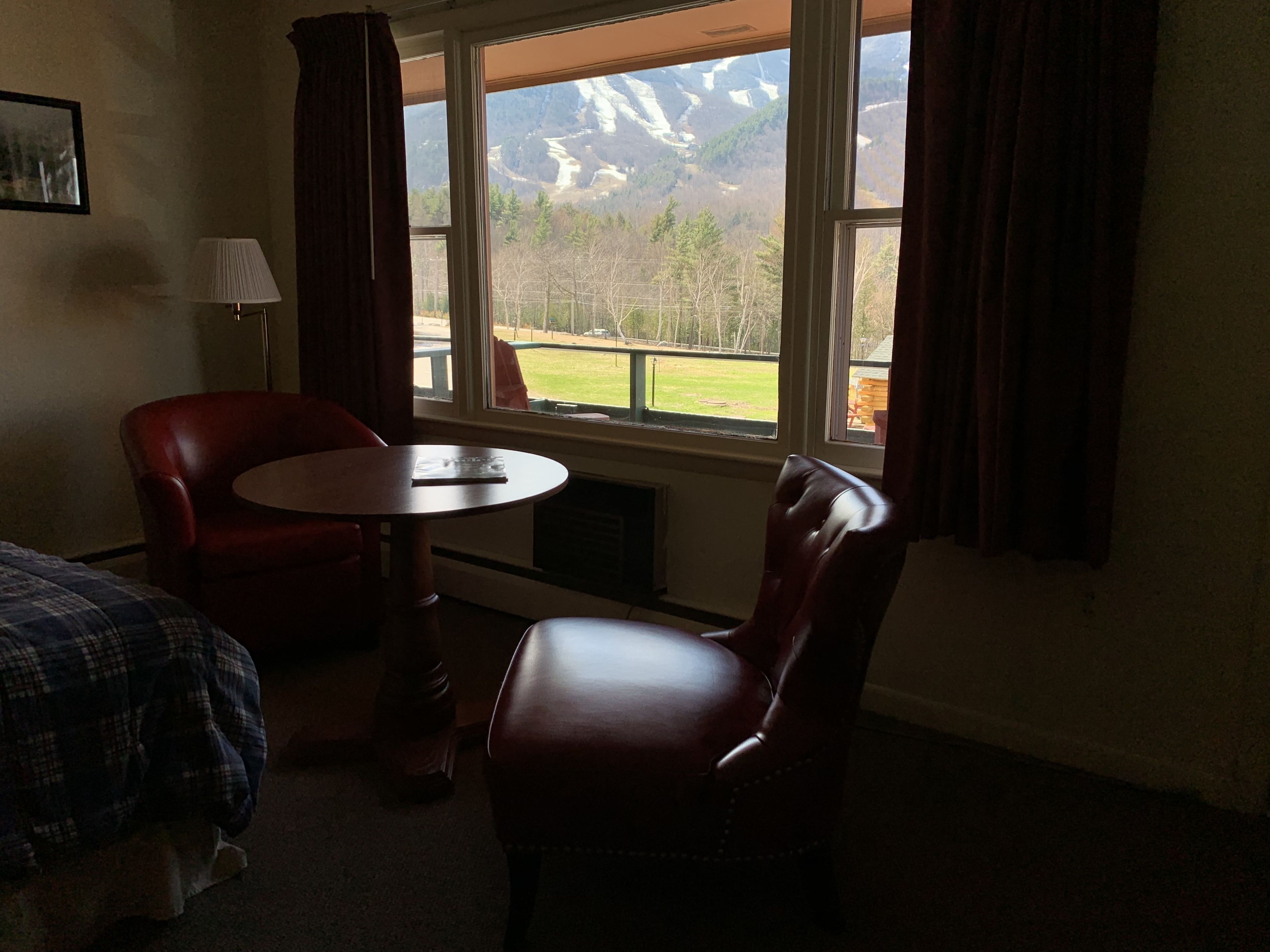 Una habitación con cama, una mesa y dos sillas. Se ve una ventana con vistas a montañas y un área verde.
