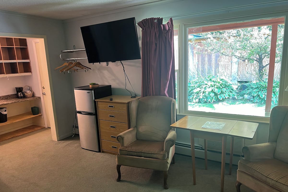 A compact living room with a sofa, a small table, a TV mounted on the wall, and a window with a view of greenery.
