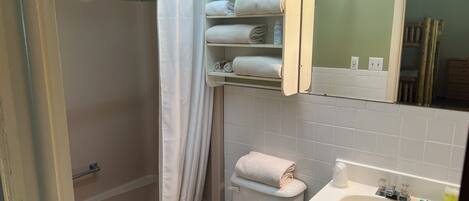 A bathroom with a shower, toilet, and sink. There are towels on the wall and a shelf with more towels.