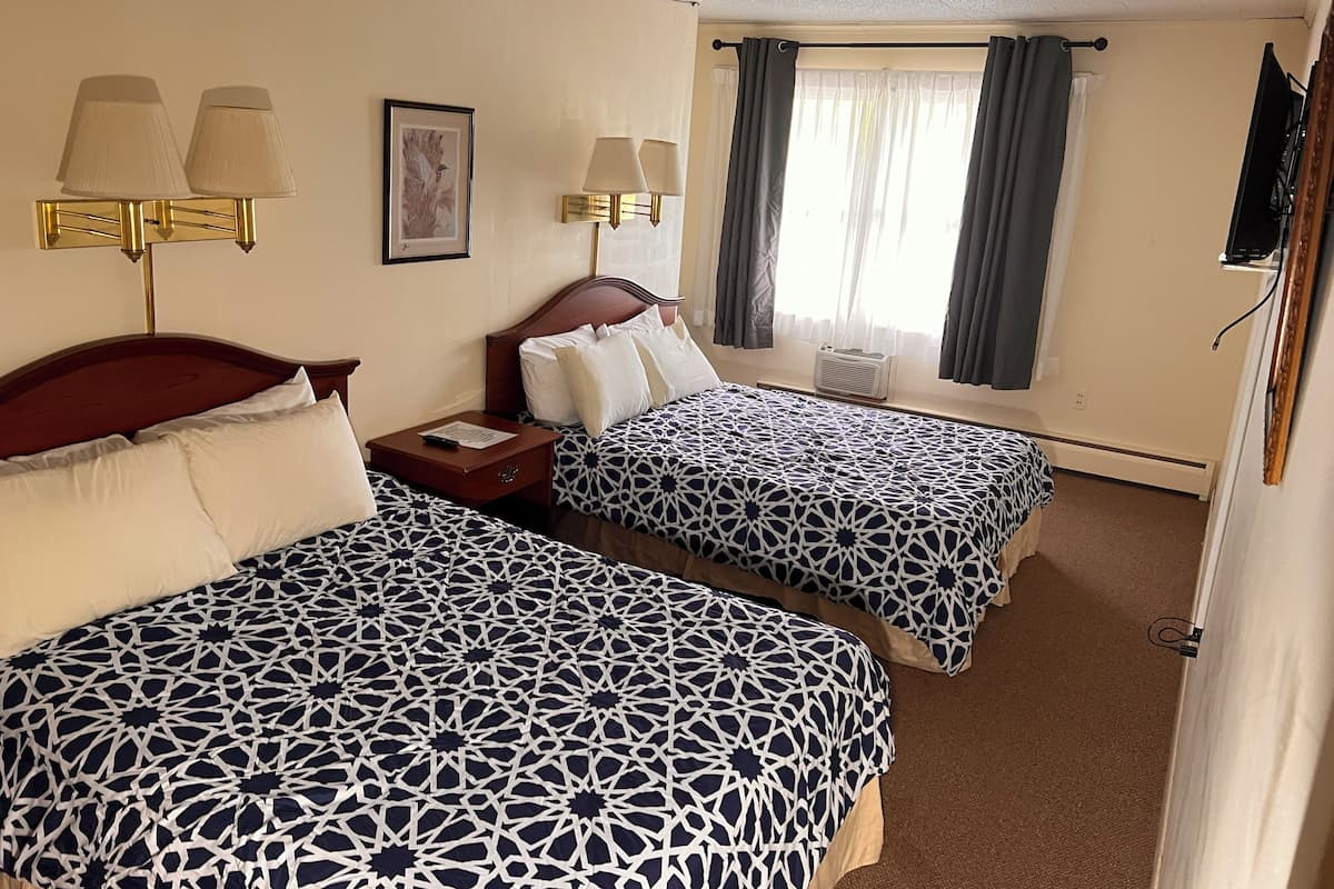 A hotel room with two beds, each with a patterned bedspread, a nightstand with a lamp, a framed picture on the wall, and a window with curtains.