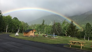 Une cabane en bois avec une table de pique-nique, située dans un parc avec une montagne en arrière-plan et un arc-en-ciel.