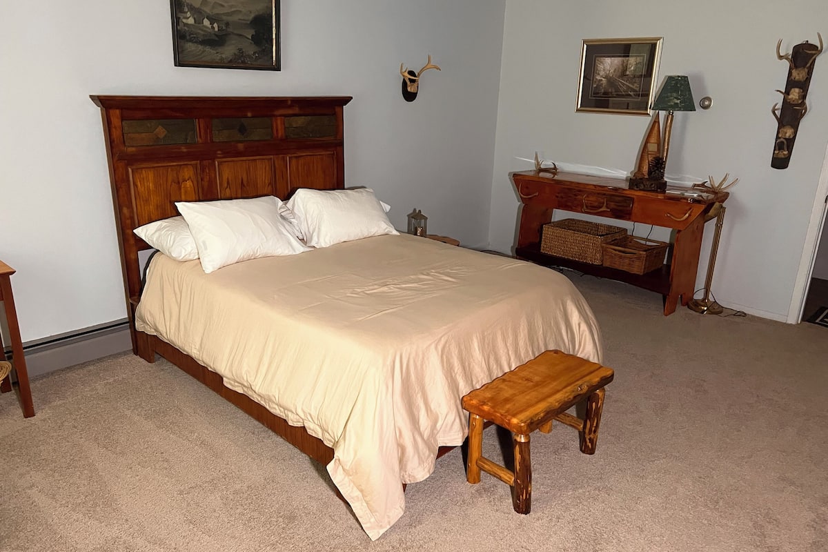 A bedroom with a wooden bed, a bedside table, a nightstand, a desk with a lamp, and wall-mounted decor.