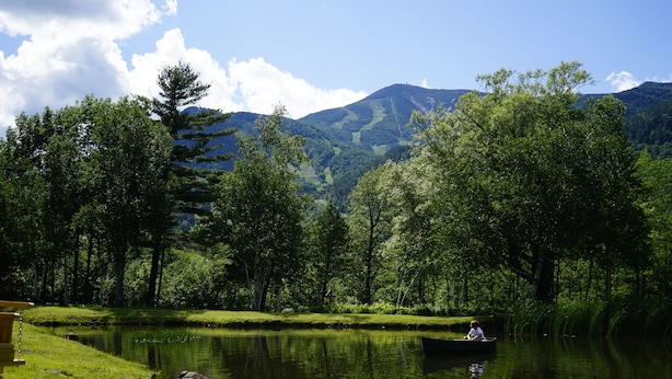 Une scène paisible au bord de la rivière, avec une personne dans un bateau, entourée d’une végétation luxuriante et des montagnes en arrière-plan.