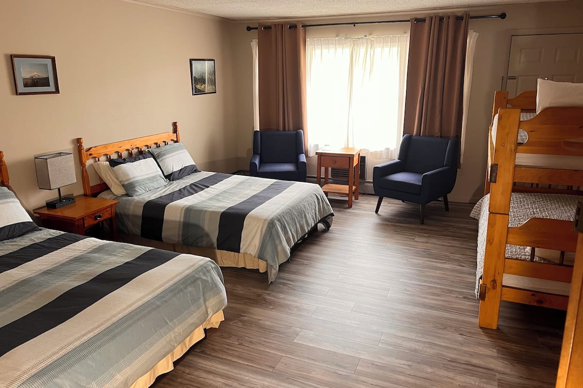 A hotel room with two beds, a wooden bunk bed, a desk, two chairs, and a window with curtains.