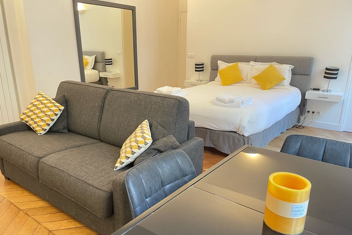 Ein modernes Hotelzimmer mit grauem Sofa, einem Bett mit weißer und gelber Bettwäsche, einem Tisch mit Kerze und einem Spiegel an der Wand.