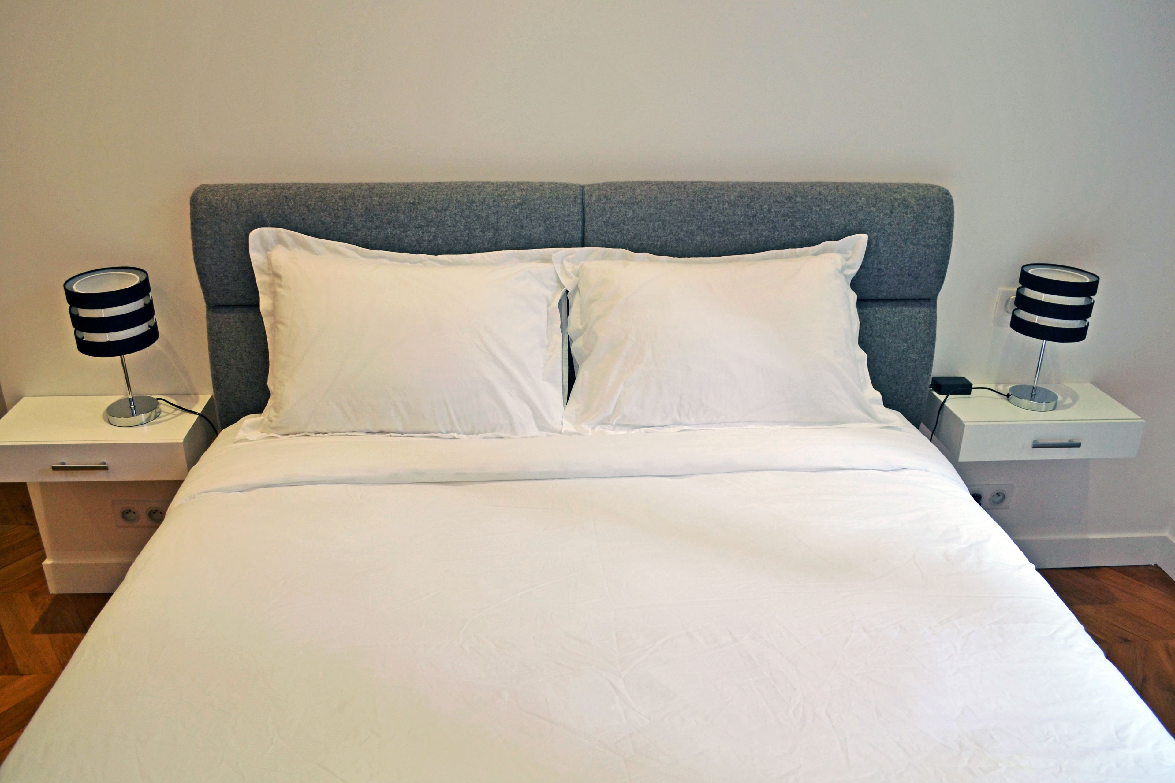 Una cama bien hecha con sábanas blancas, un cabecero tapizado en gris y dos mesitas de noche con lámparas blanco y negro.