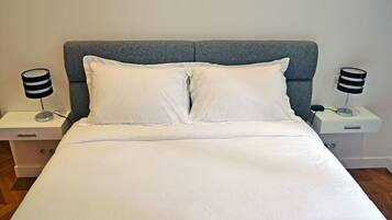 Una cama bien hecha con sábanas blancas, un cabecero tapizado en gris y dos mesitas de noche con lámparas blanco y negro.