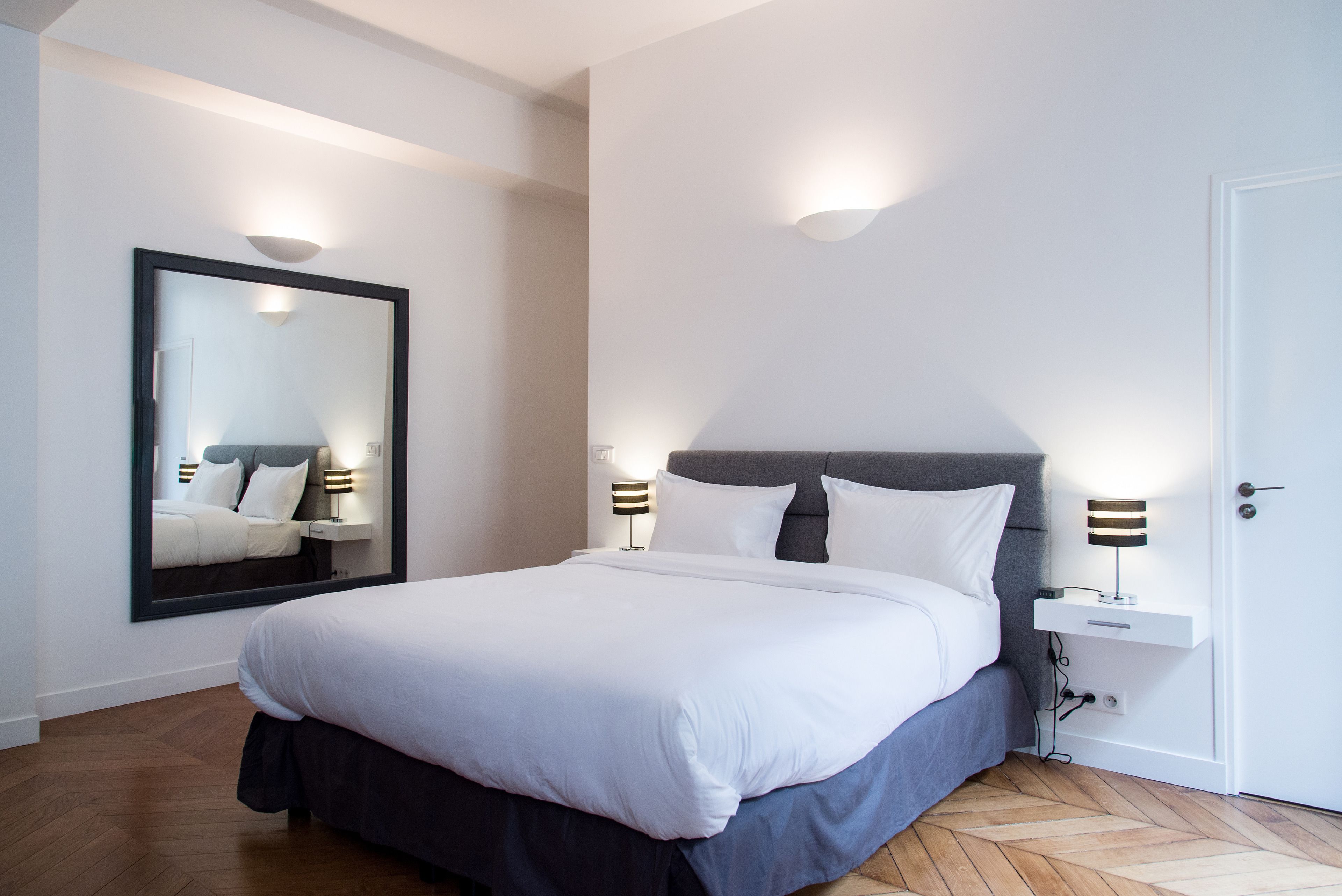 A modern hotel room with a large bed, two bedside tables with lamps, a large mirror, and a door.