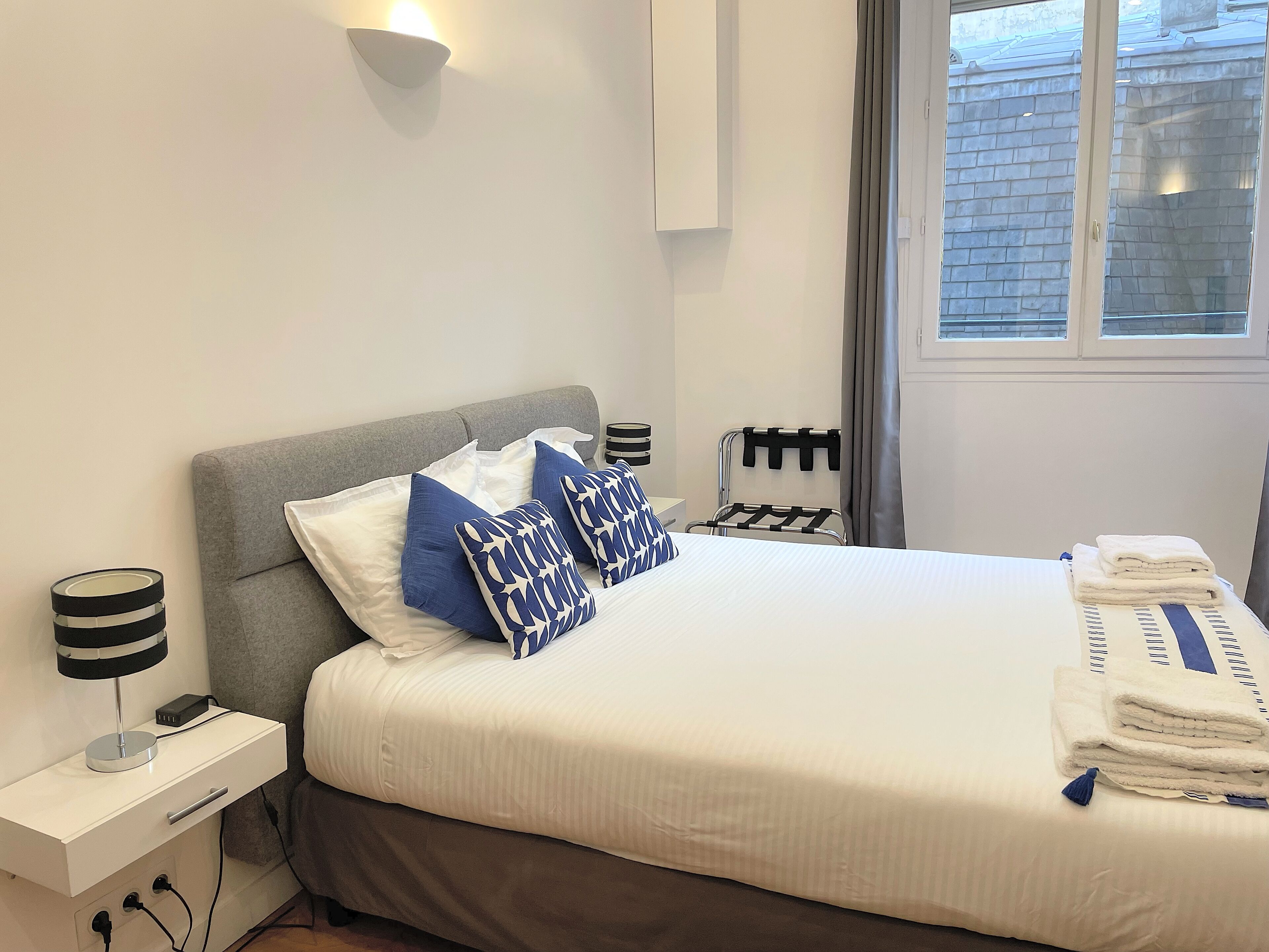 A neatly made bed with white and blue pillows, a grey headboard, and a bedside table with a lamp and a power outlet.