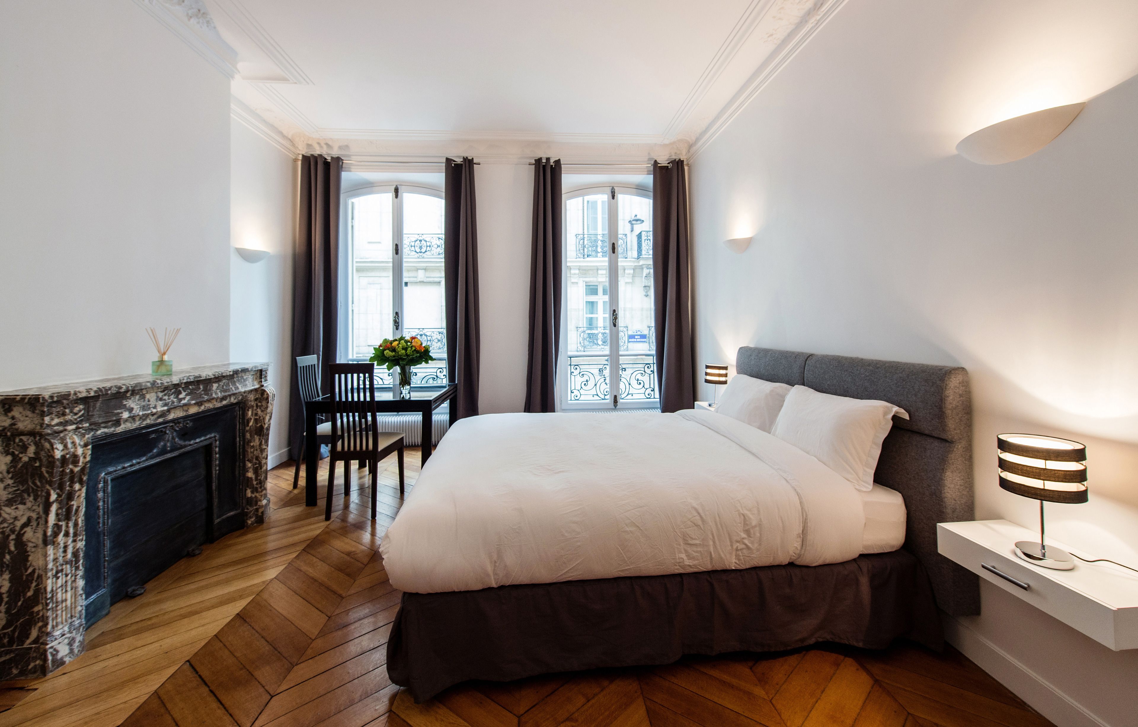 Une chambre d’hôtel avec un grand lit, une cheminée, une table à manger avec des chaises et une vue sur la ville par les fenêtres.