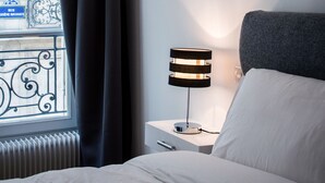 A hotel room with a bed, a bedside table with a lamp, a window with curtains, and a view of a building with balconies. - Paris Square (Paris)