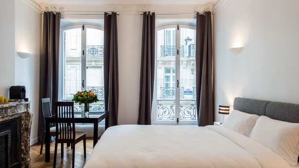 A hotel room with a large bed, a desk with a chair, a vase of flowers, and a view of the city through the windows. - Paris Square (Paris)