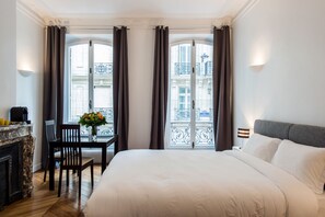Habitación de hotel con una cama grande, un escritorio con una silla, un florero y vistas de la ciudad a través de las ventanas.