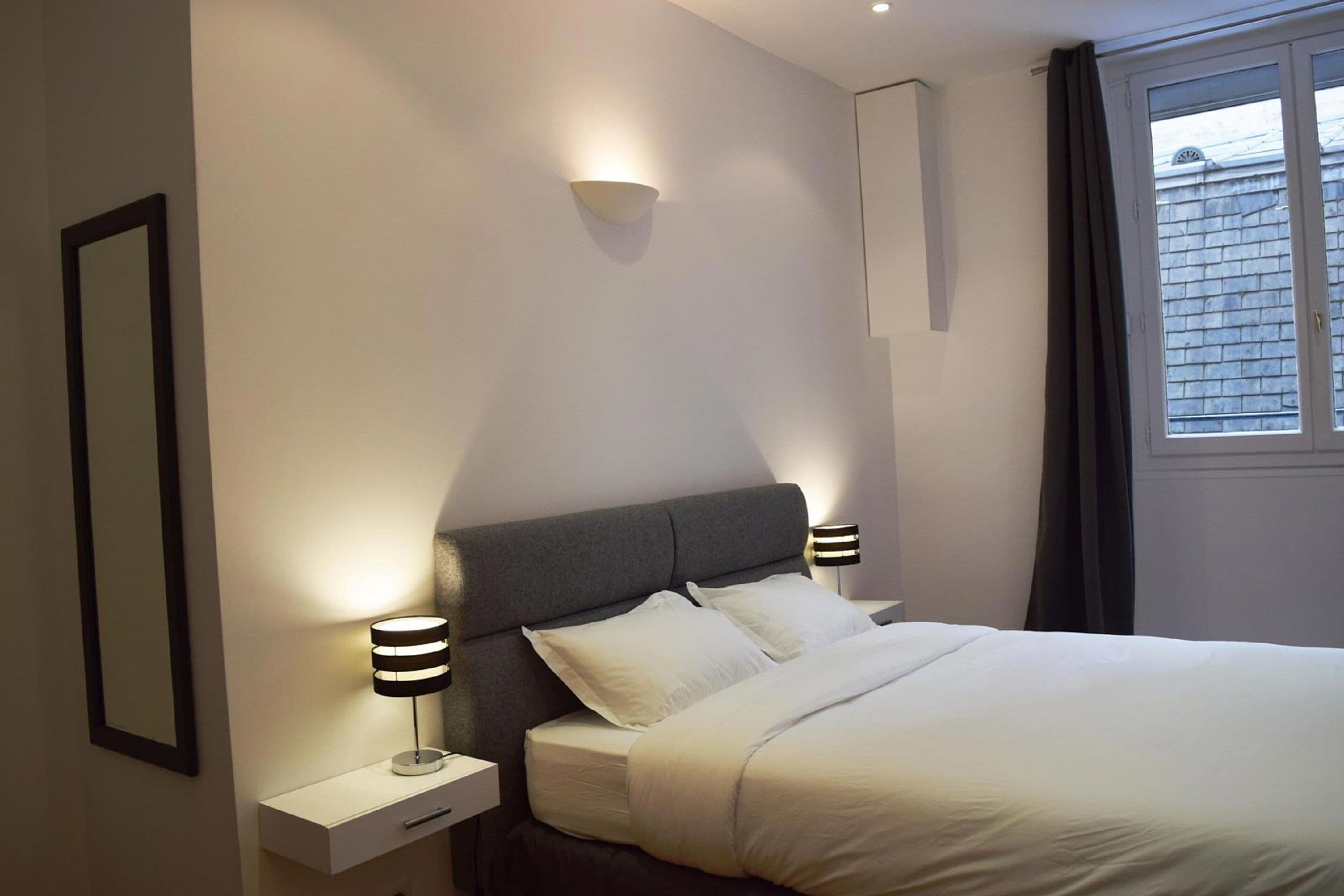A modern bedroom with a large bed, bedside lamps, a nightstand, and a window with curtains.