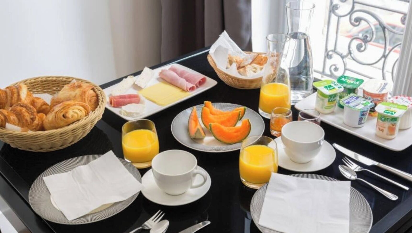 A breakfast tray with croissants, ham, cheese, and melon on a balcony table with breakfast drinks and yogurt containers.