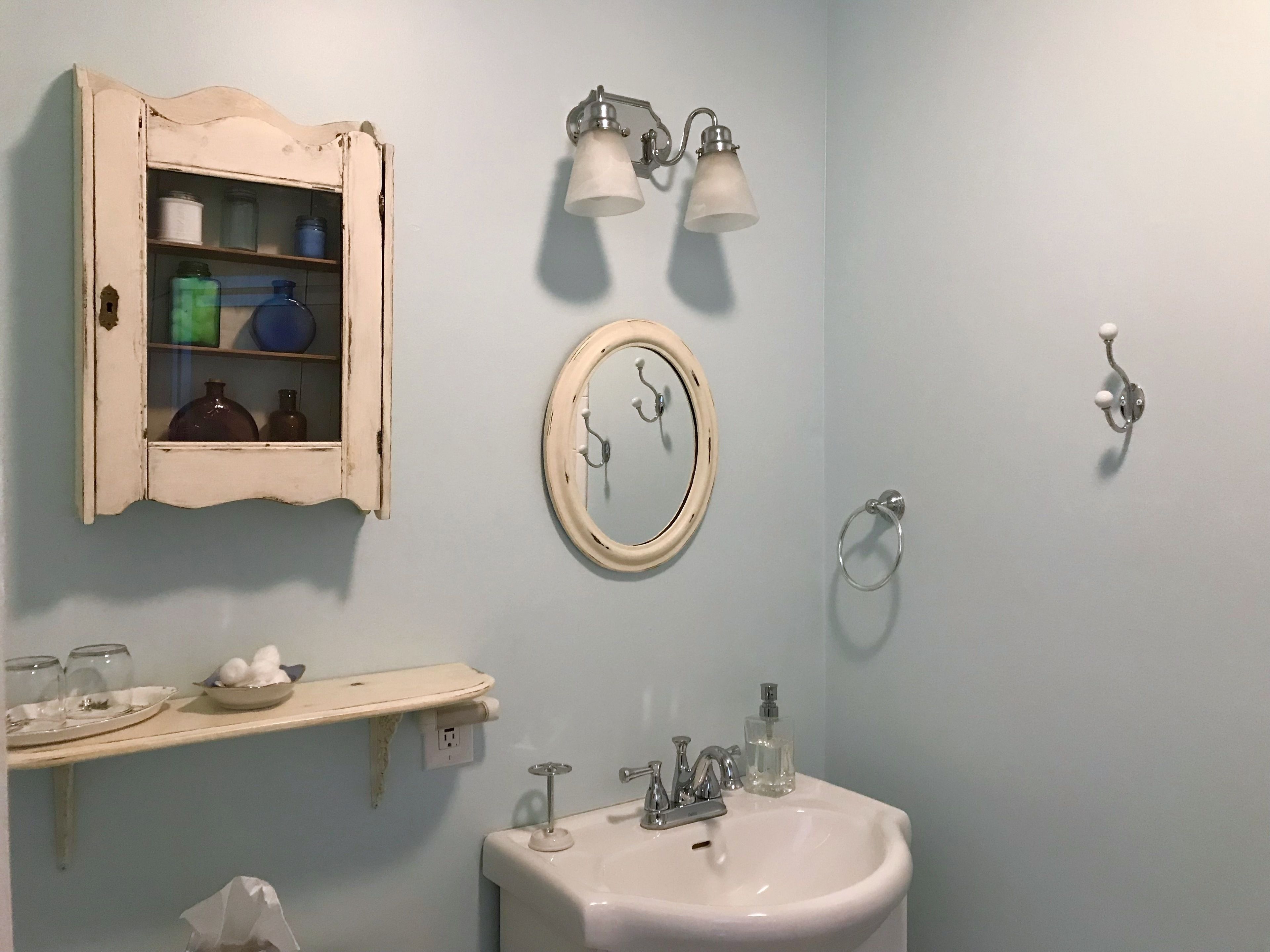 Un baño con lavamanos blanco, un espejo, un armario con frascos y un tozaleto.