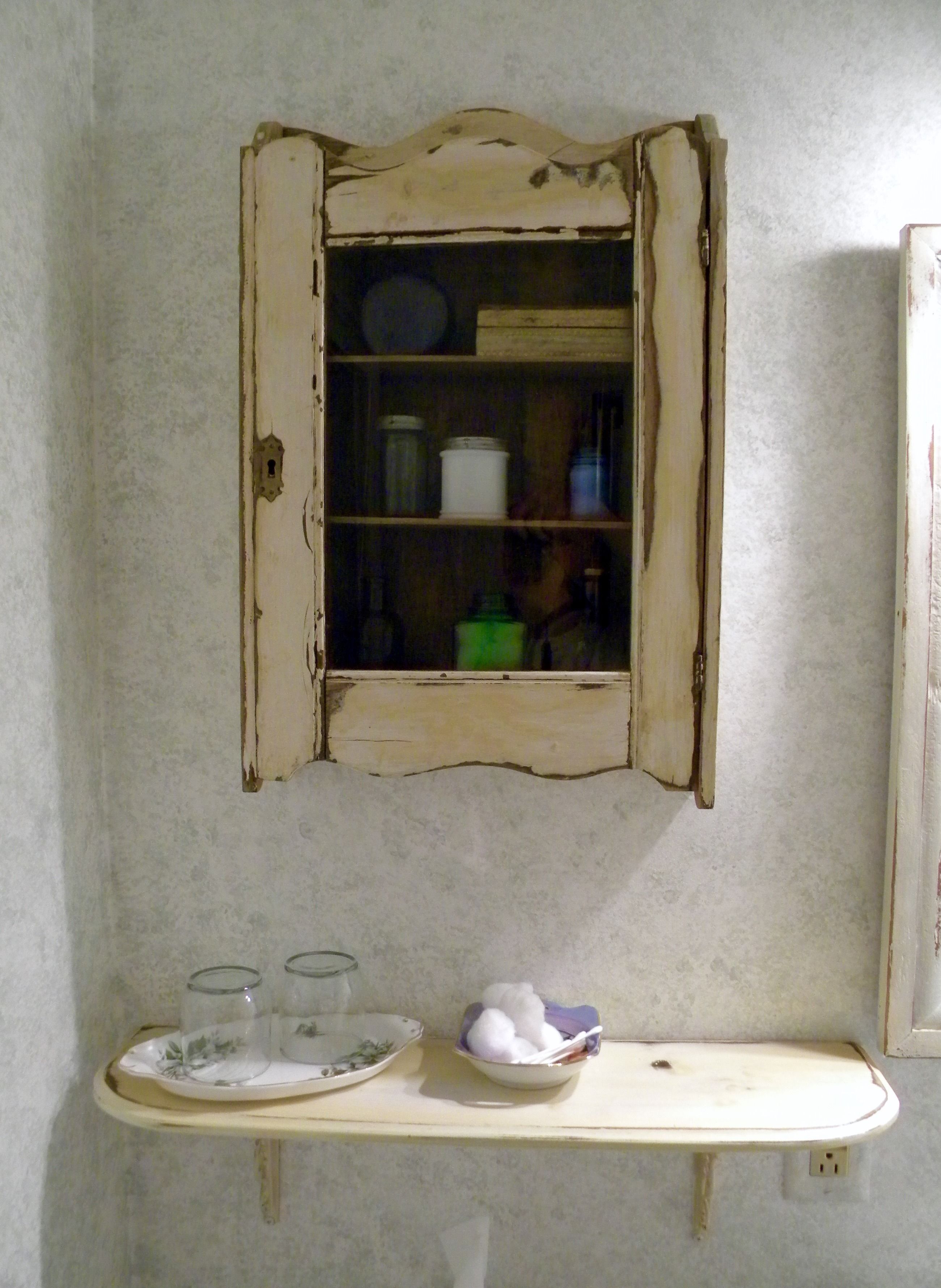 A rustic kitchen with a wooden cabinet containing various kitchen items and a small shelf below holding glasses and a bowl.
