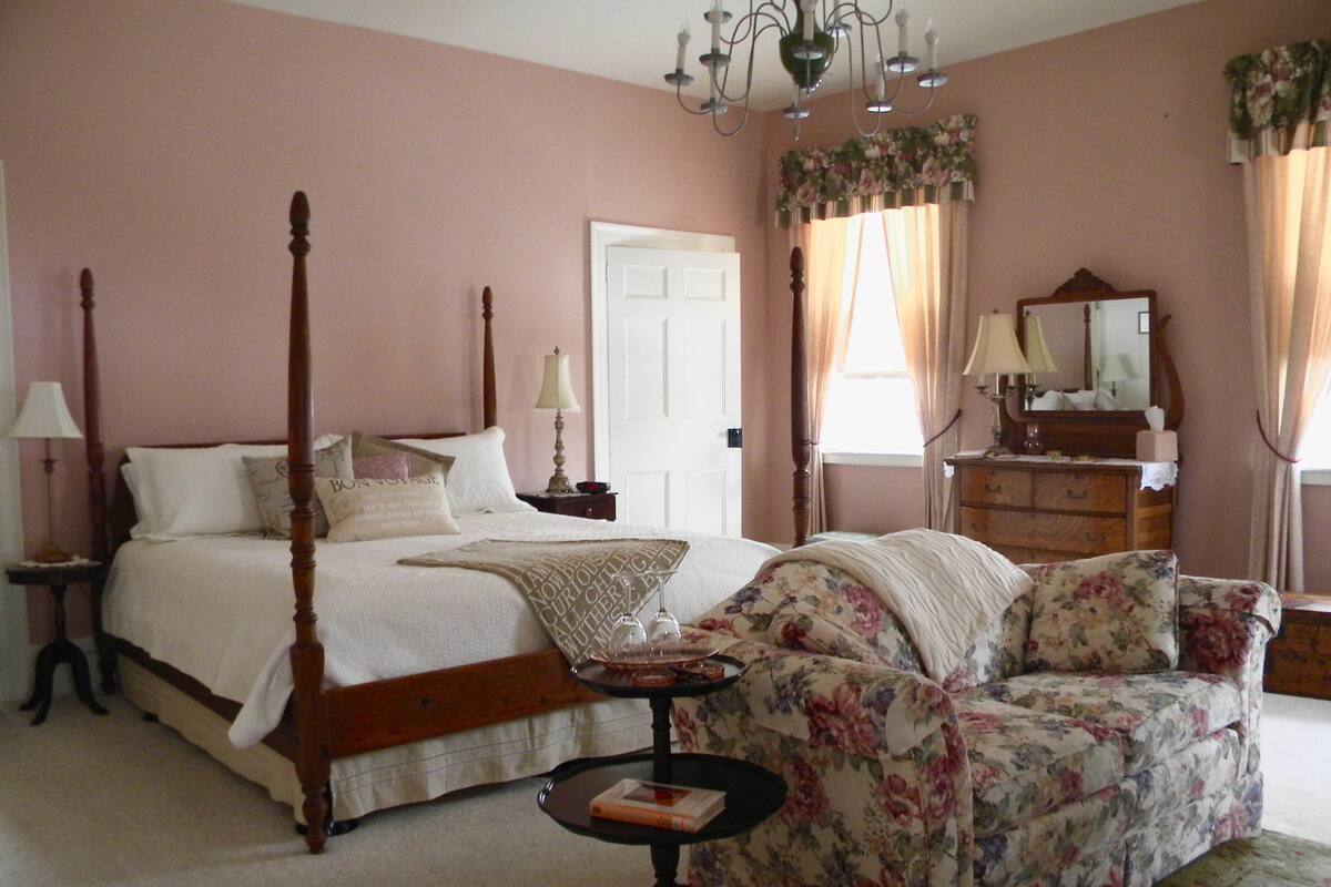 A bedroom with a four-poster bed, a floral sofa, a small round table, and a dresser with a mirror.