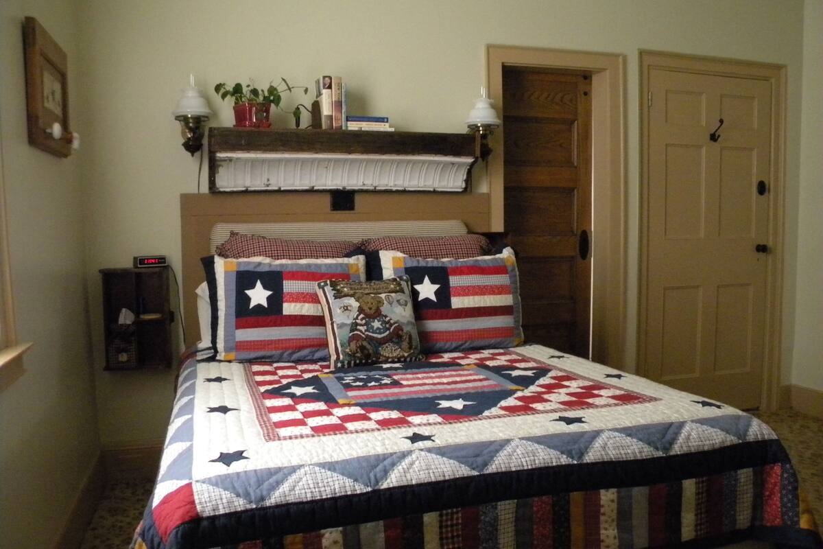 A bedroom with a bed featuring a quilt with a star pattern, a wooden headboard, a nightstand with a clock, and a door to the right.