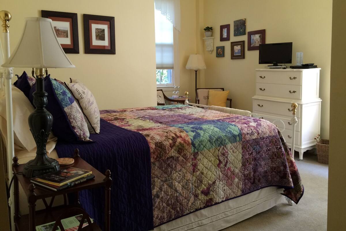 A bedroom with a bed, a nightstand, a lamp, a dresser, and several framed pictures on the wall.