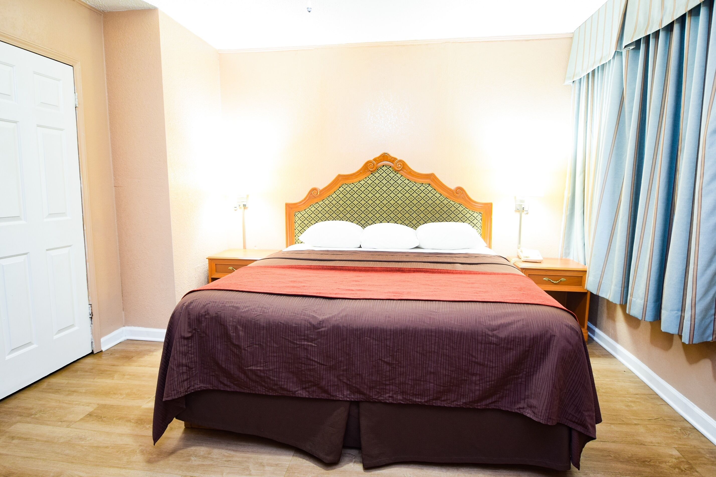 Una cama bien hecha con cabecera de madera, una mesita de noche con lámpara y una ventana con cortinas.