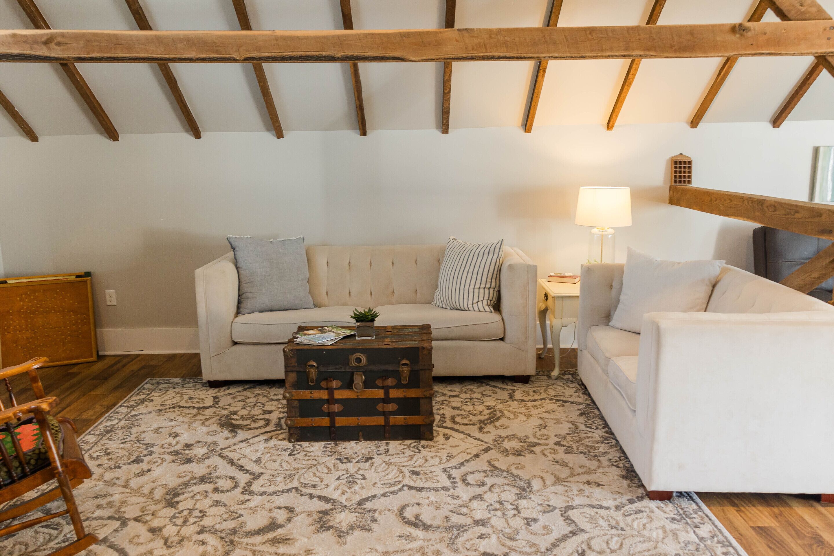 Una sala de estar acogedora con un alto techo de madera, un sofá color beige, un sillón blanco, un tapete estampado y un aparador de madera.
