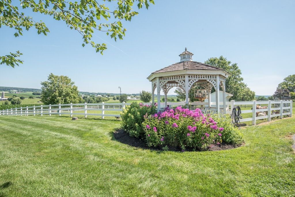 A gazebo with a white lattice roof and a small tower, surrounded by a white fence and vibrant pink flowers.