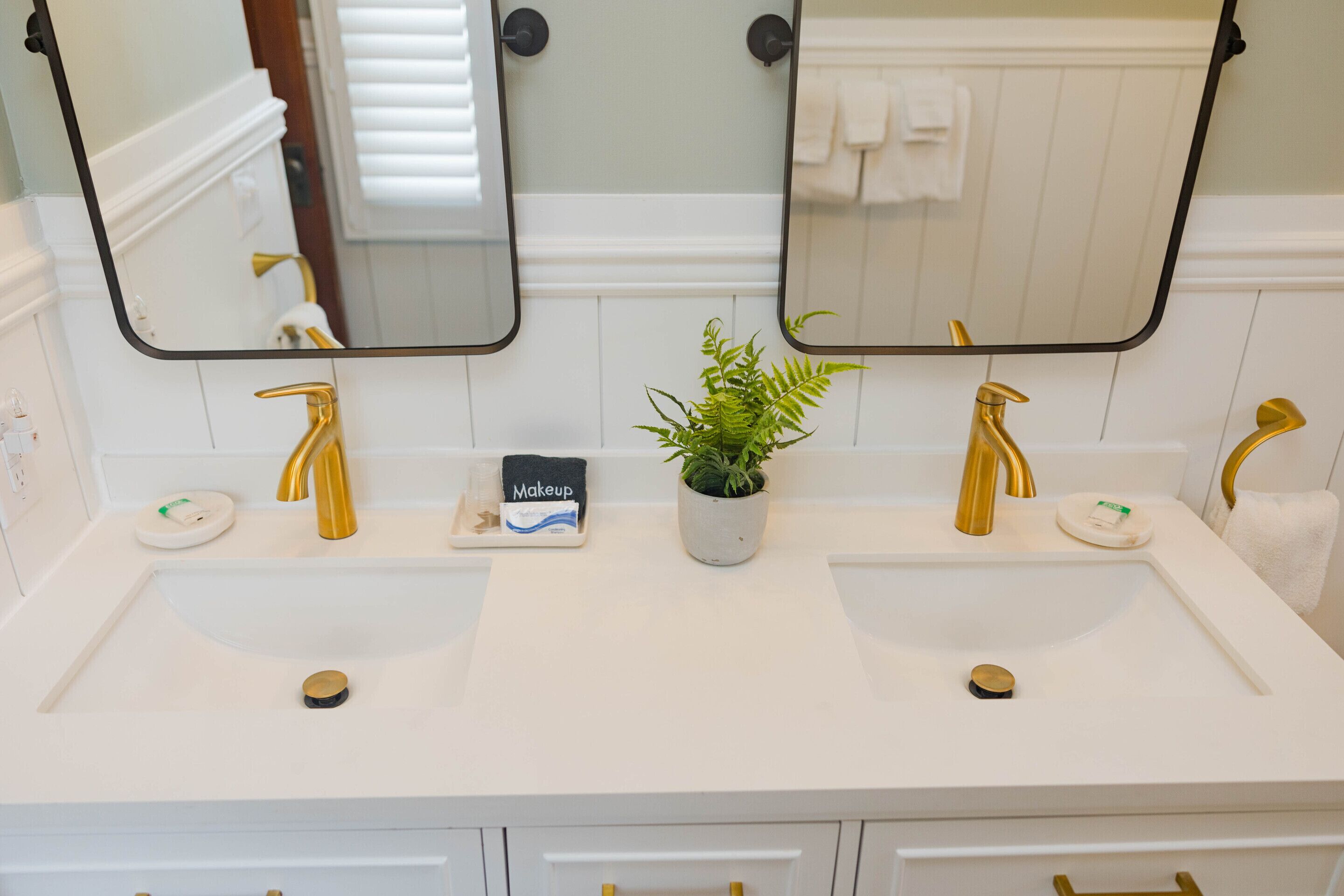 A bathroom with two sinks, each with a gold faucet, a potted plant, and a makeup station.