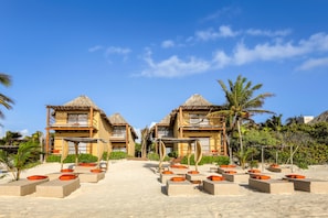 A resort with two thatched-roof buildings, outdoor seating areas with orange cushions, and a clear blue sky.