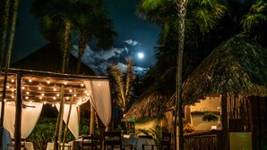 Un espace piscine aménagé pour la nuit, avec un coin salon couvert, des palmiers et une structure dotée d’un toit de chaume.