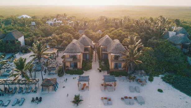 Un complexe hôtelier doté de villas aux toits de chaume, d’une plage de sable et de palmiers.
