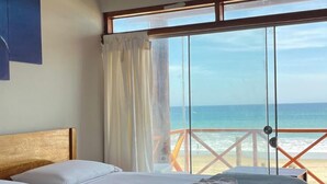 A bedroom with a bed, a ceiling fan, a large window with a view of the beach, and a balcony with a railing.
