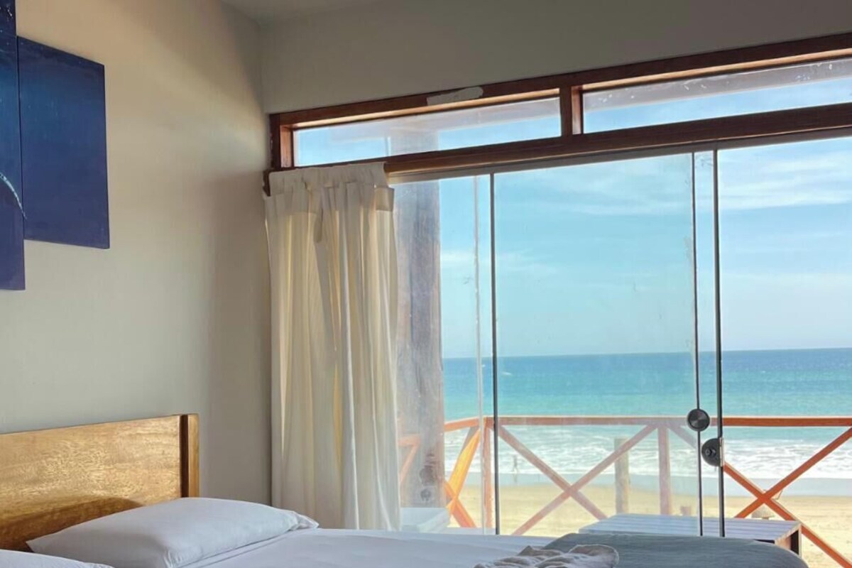 A bedroom with a bed, a ceiling fan, a large window with a view of the beach, and a balcony with a railing.