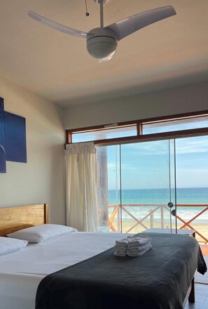 A bedroom with a bed, a ceiling fan, a large window with a view of the beach, and a balcony with a railing.