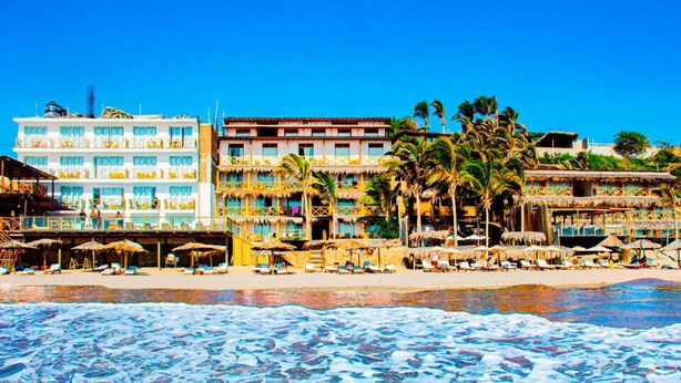 A beachfront hotel with multiple buildings, including a large hotel with balconies and a smaller structure with thatched roofs.
