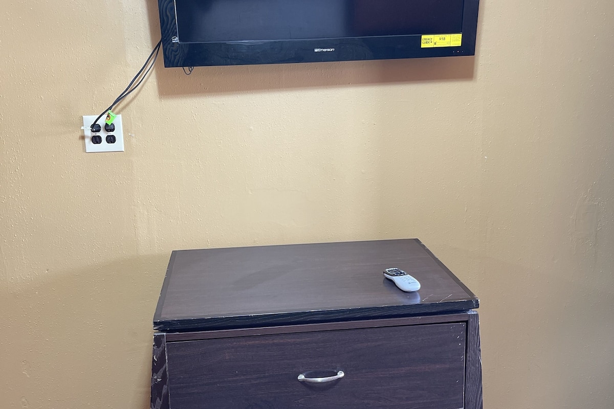 Un téléviseur à écran plat fixé à un mur de couleur beige, un meuble en bois foncé avec des tiroirs, et une télécommande posée sur le dessus.