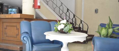 Un vestíbulo con sillones azules, una mesa blanca con un arreglo floral, un extintor y una escalera.