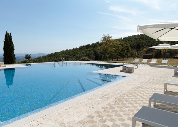 Una piscina con agua cristalina azul, rodeada por una terraza con mosaicos, sillones de descanso y sombrillas.