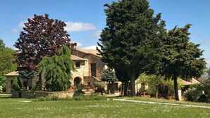 Un edificio de piedra con techo de tejas, rodeado de un jardín cuidado con diversas especies de árboles y césped.
