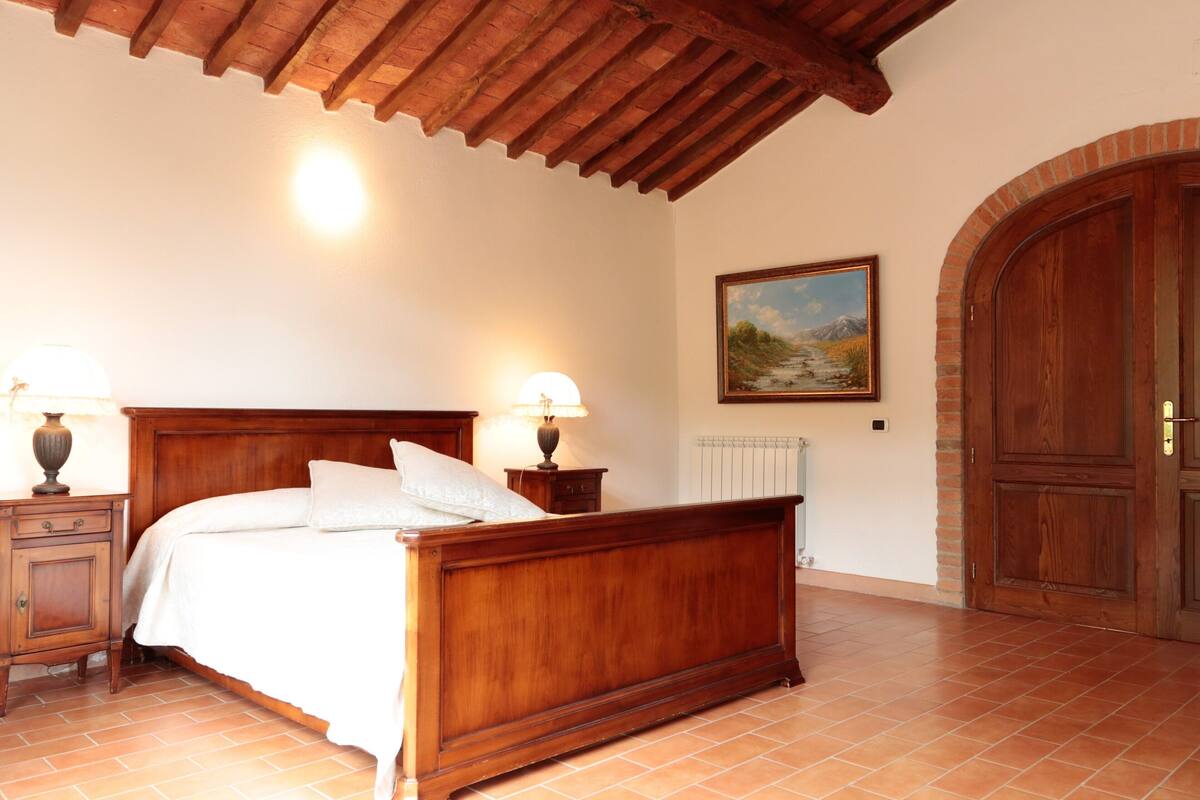 Une chambre avec un lit en bois, deux tables de chevet avec des lampes, un radiateur, un tableau au mur et une grande porte en bois.