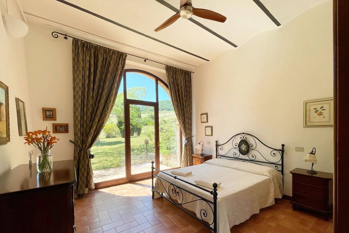 Une chambre à coucher comprenant un lit, un ventilateur de plafond, une fenêtre avec des rideaux, une table de chevet et une commode.