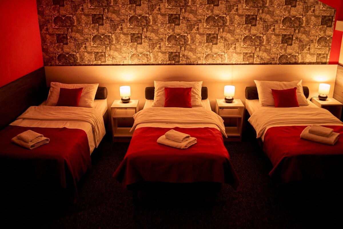 Une chambre d’hôtel avec trois lits simples, chacun recouvert d’une couverture rouge et agrémenté d’oreillers blancs, et un mur orné d’un motif décoratif.
